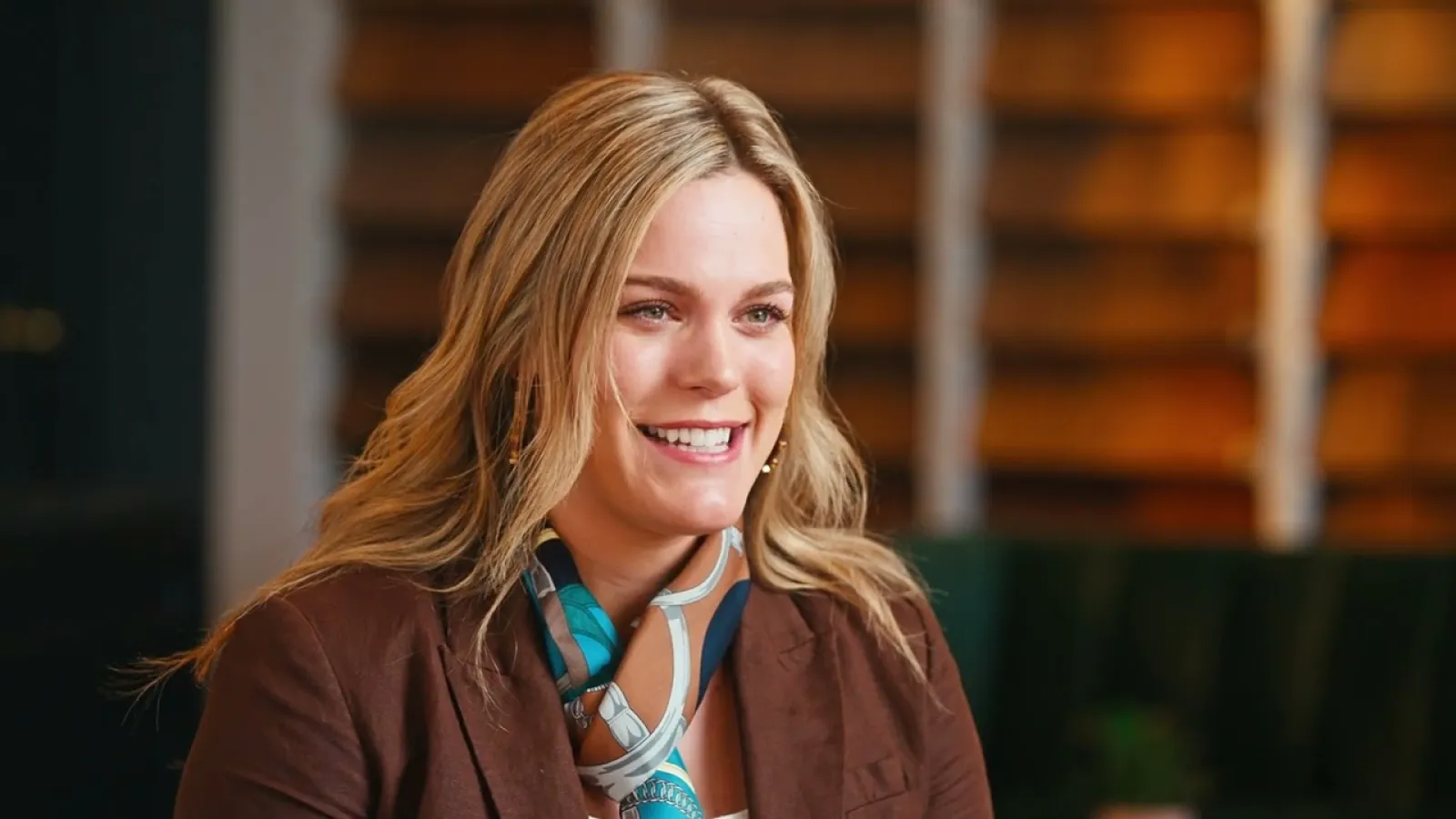 Smiling blonde woman wearing a brown blazer and patterned scarf in a warmly lit indoor setting