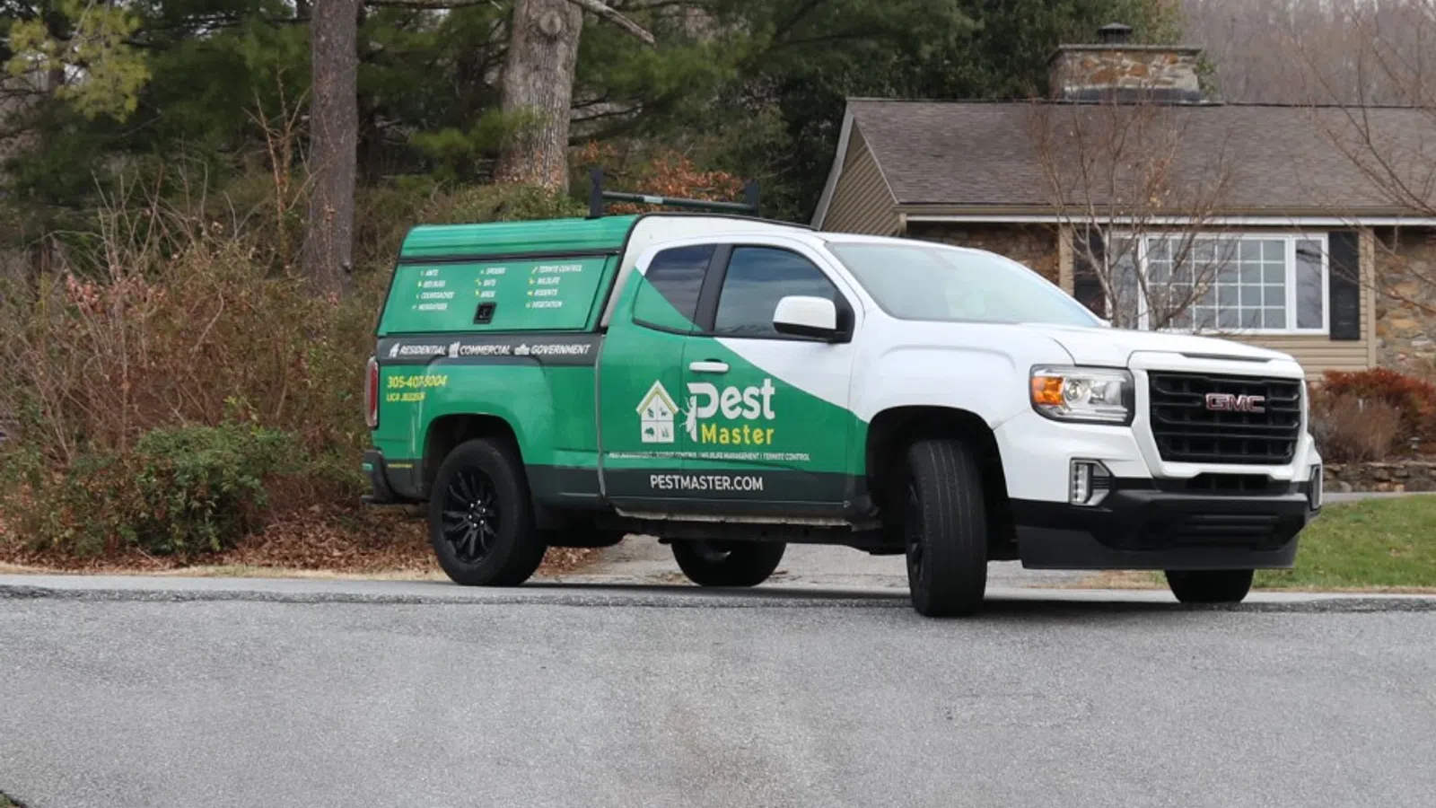 Green and white pest control truck parked on a driveway in front of suburban houses with trees and lawns.