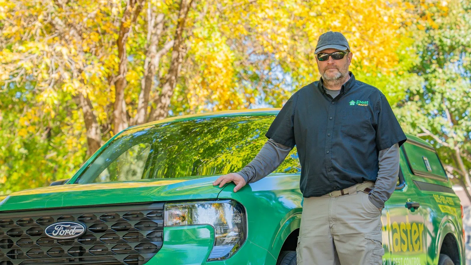 Man wearing workwear standing next to a green Ford pickup truck in a park with autumn trees in background.
