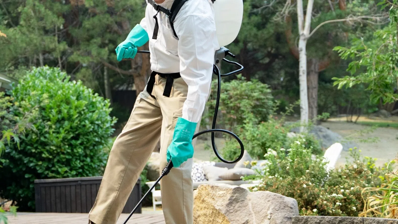 Man wearing protective gloves and goggles spraying pesticide outdoors near wooden deck and garden plants.