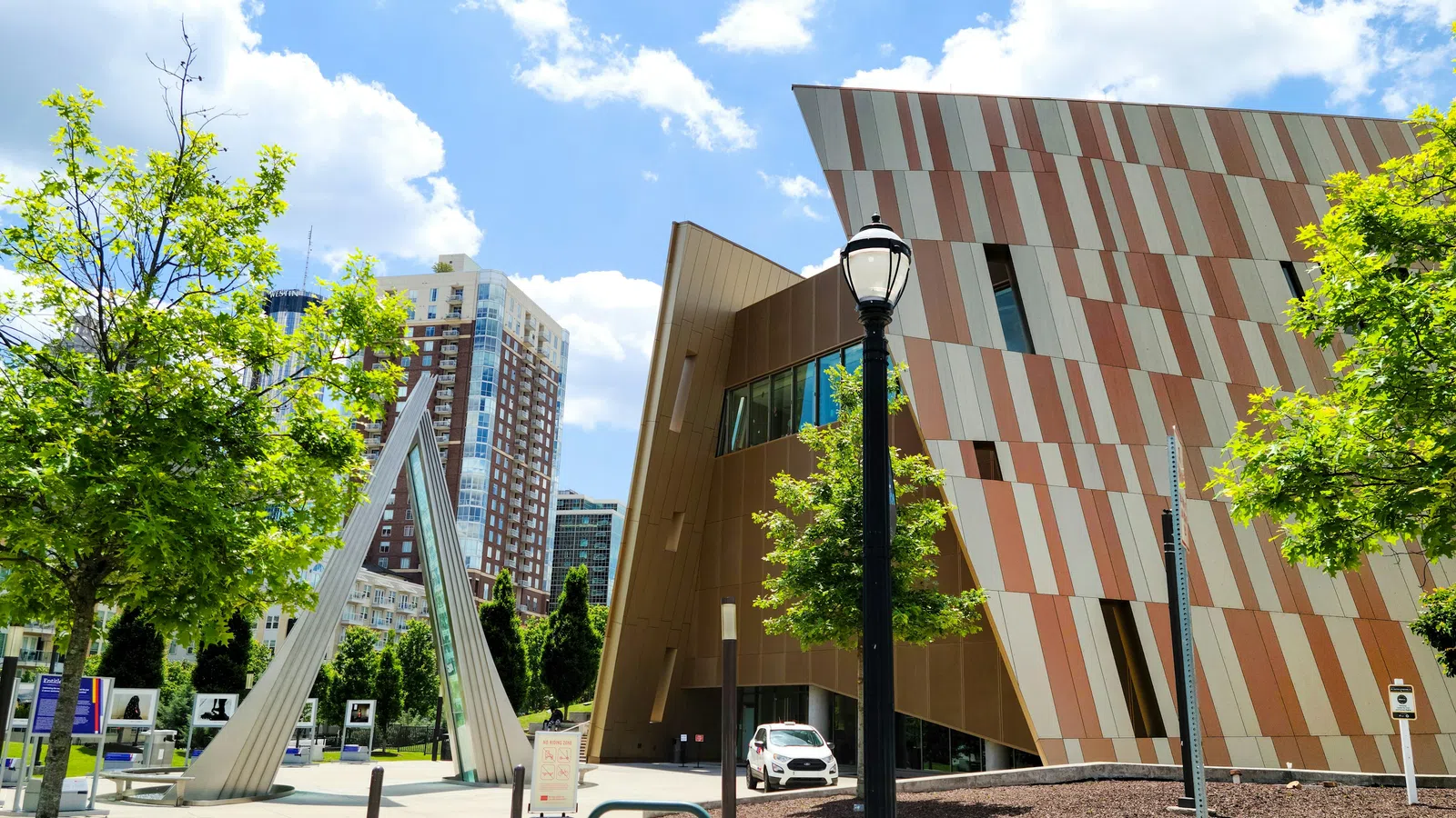 Modern building with geometric design, urban park, trees, and blue sky with clouds in the background.