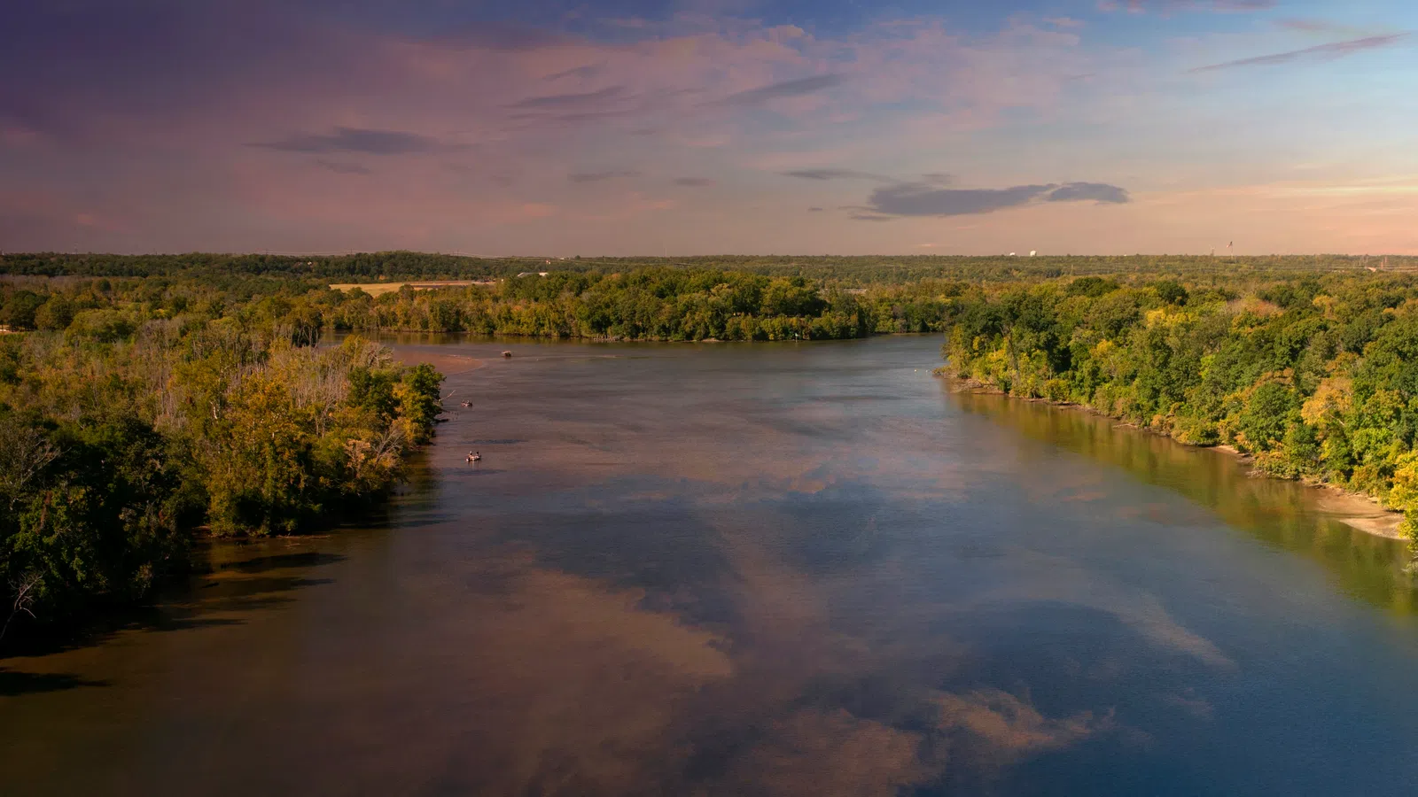 Wide river flowing through dense green forest under a partly cloudy sky at sunset or sunrise.