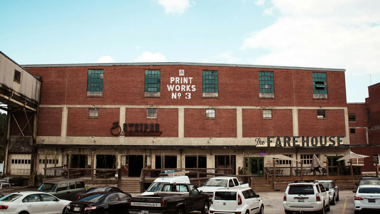 Brick industrial building with vintage signage and parked cars under a bright blue sky with clouds.