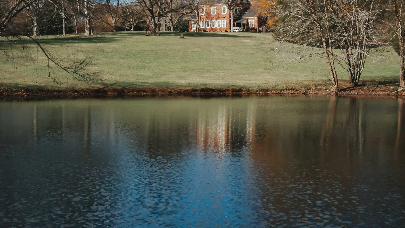 Historic brick house on green lawn with autumn trees reflected in calm pond under blue sky