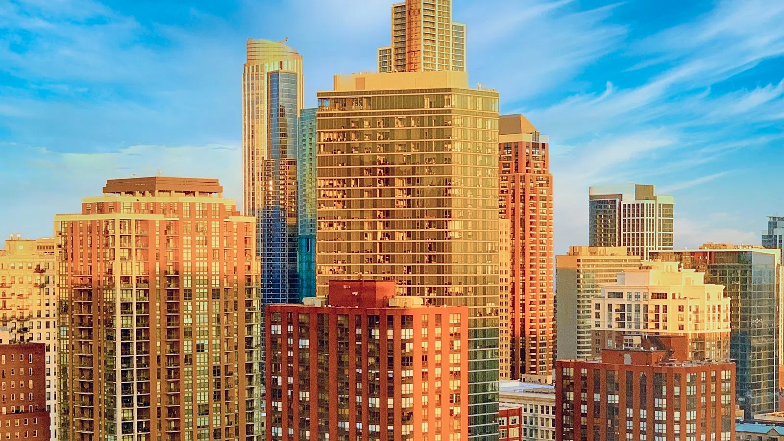 Sunlit modern high-rise buildings and green trees under a bright blue sky in a cityscape view.