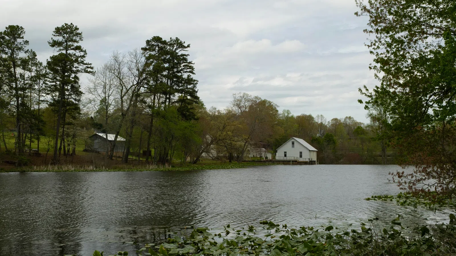 Calm lake surrounded by trees with a white house and other buildings near the water under a cloudy sky