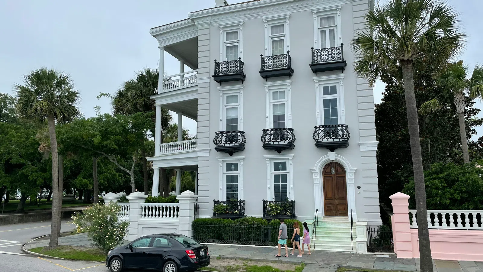 Elegant white three-story historic house with black wrought iron balconies and palm trees on a cloudy day.