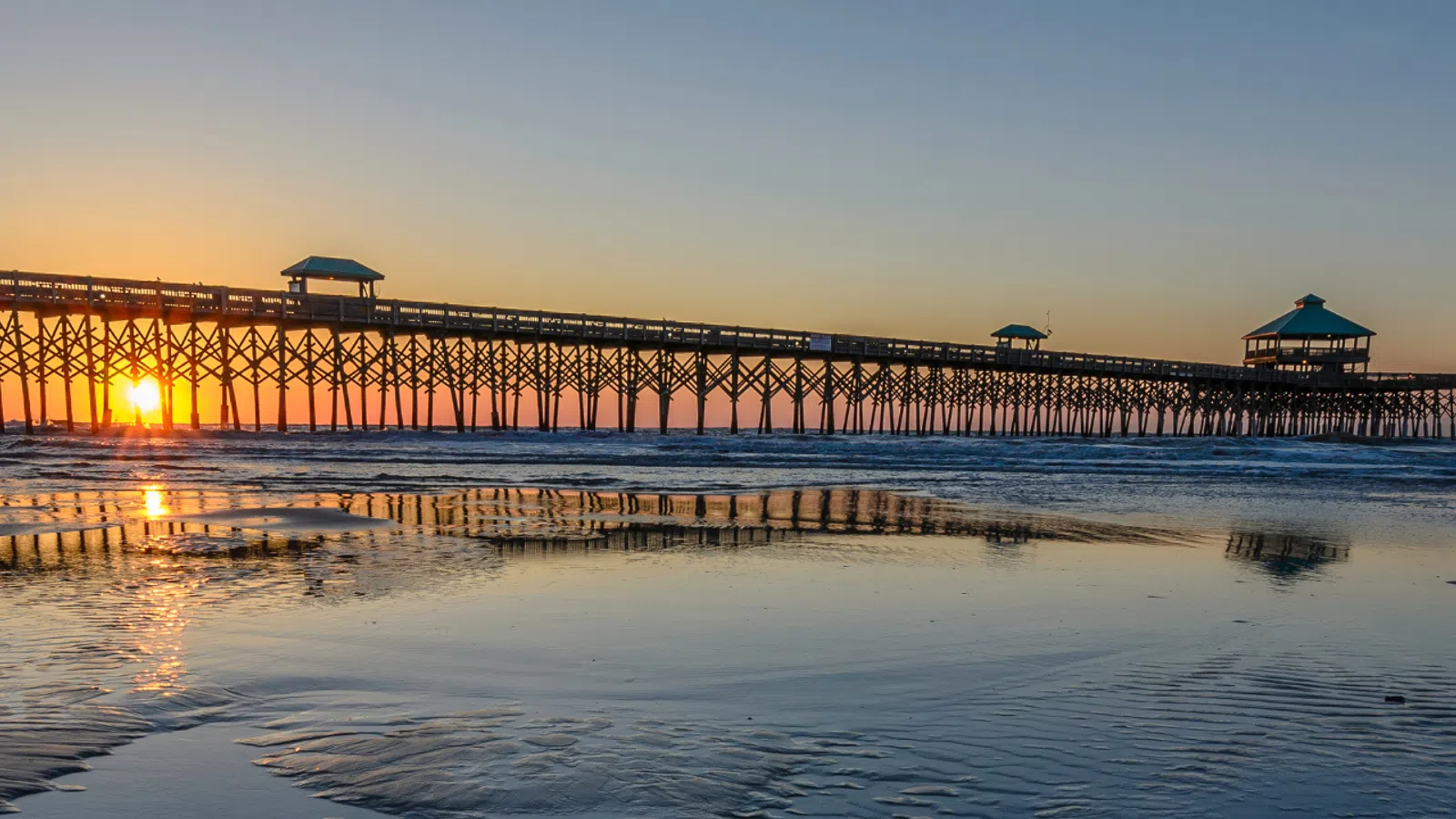 Sunset over a long wooden pier with reflections on wet sandy beach and calm ocean in the background