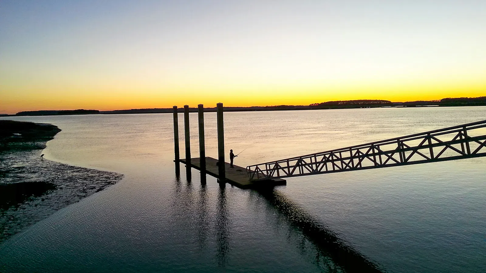 Silhouette of a person fishing on a dock at sunset with calm water and clear sky.