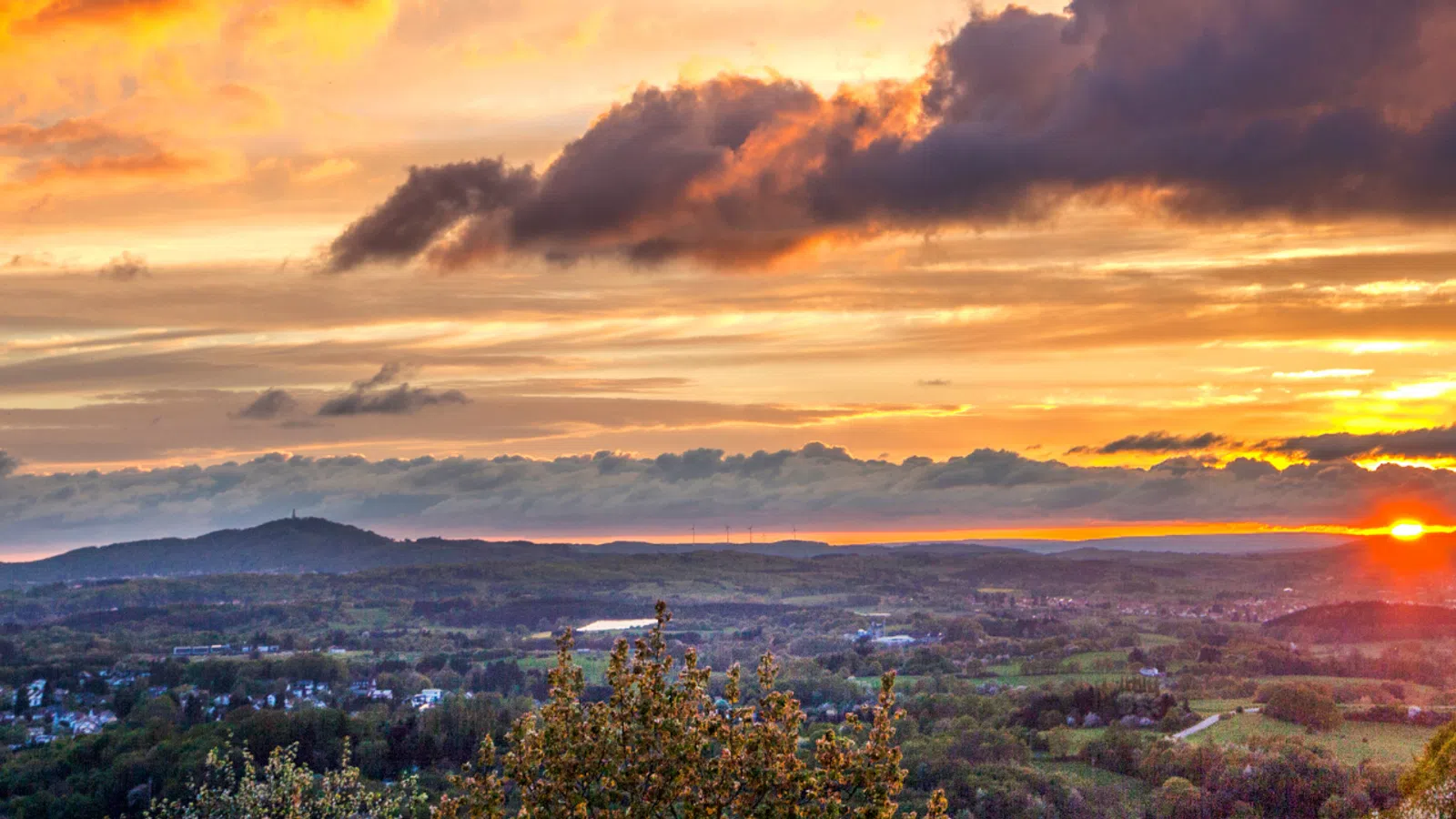 Vibrant sunset over a sprawling landscape with hills, trees, and scattered clouds illuminating the sky.