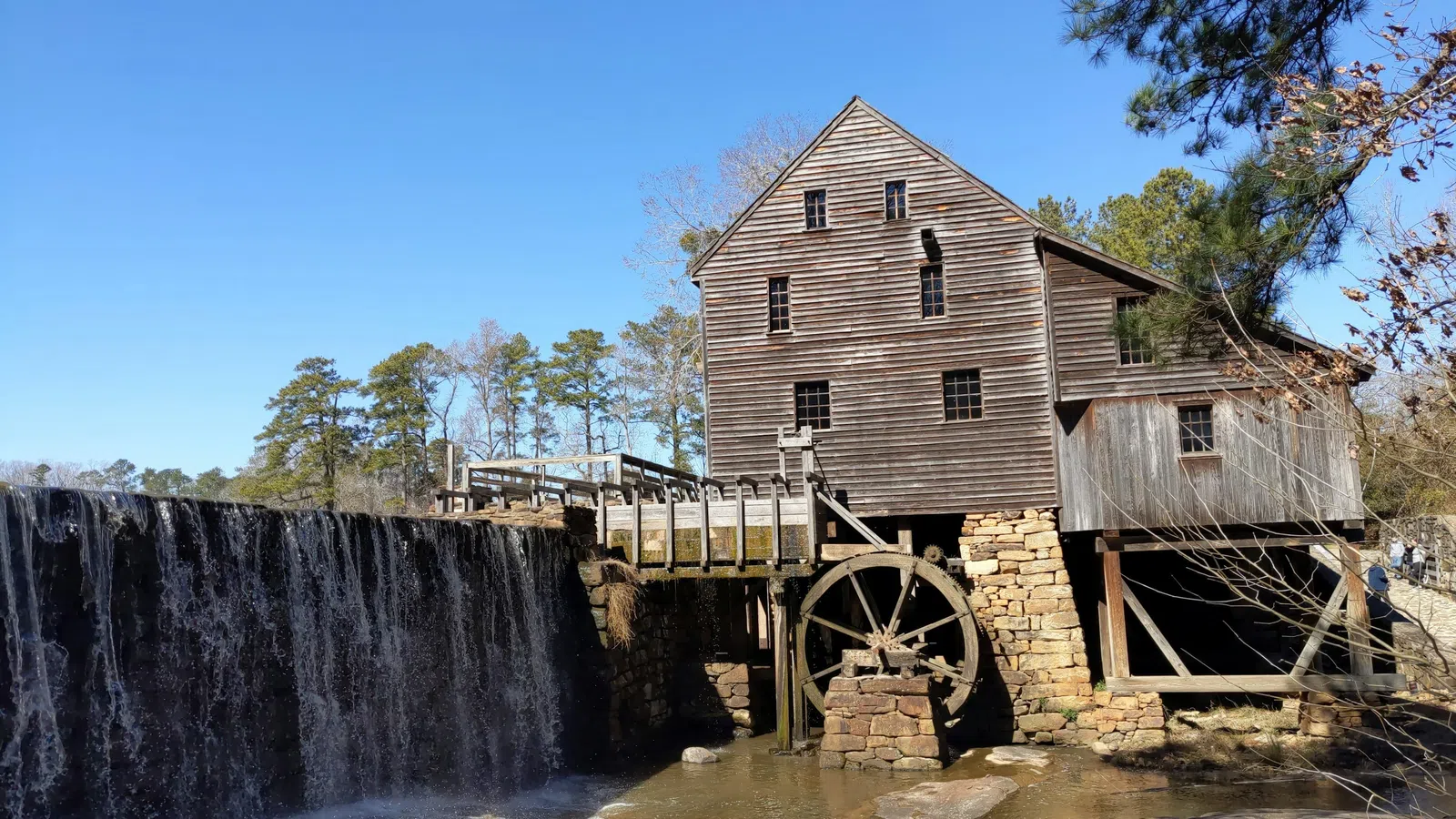 Historic wooden watermill with large wheel beside cascading waterfall under clear blue sky.