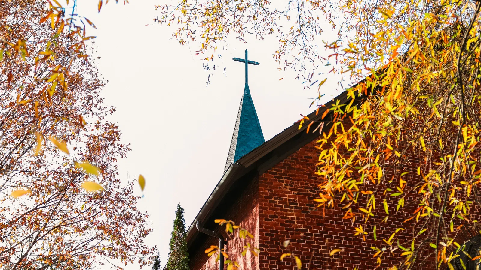 Autumn leaves frame a brick church with a cross-topped steeple against a pale sky.