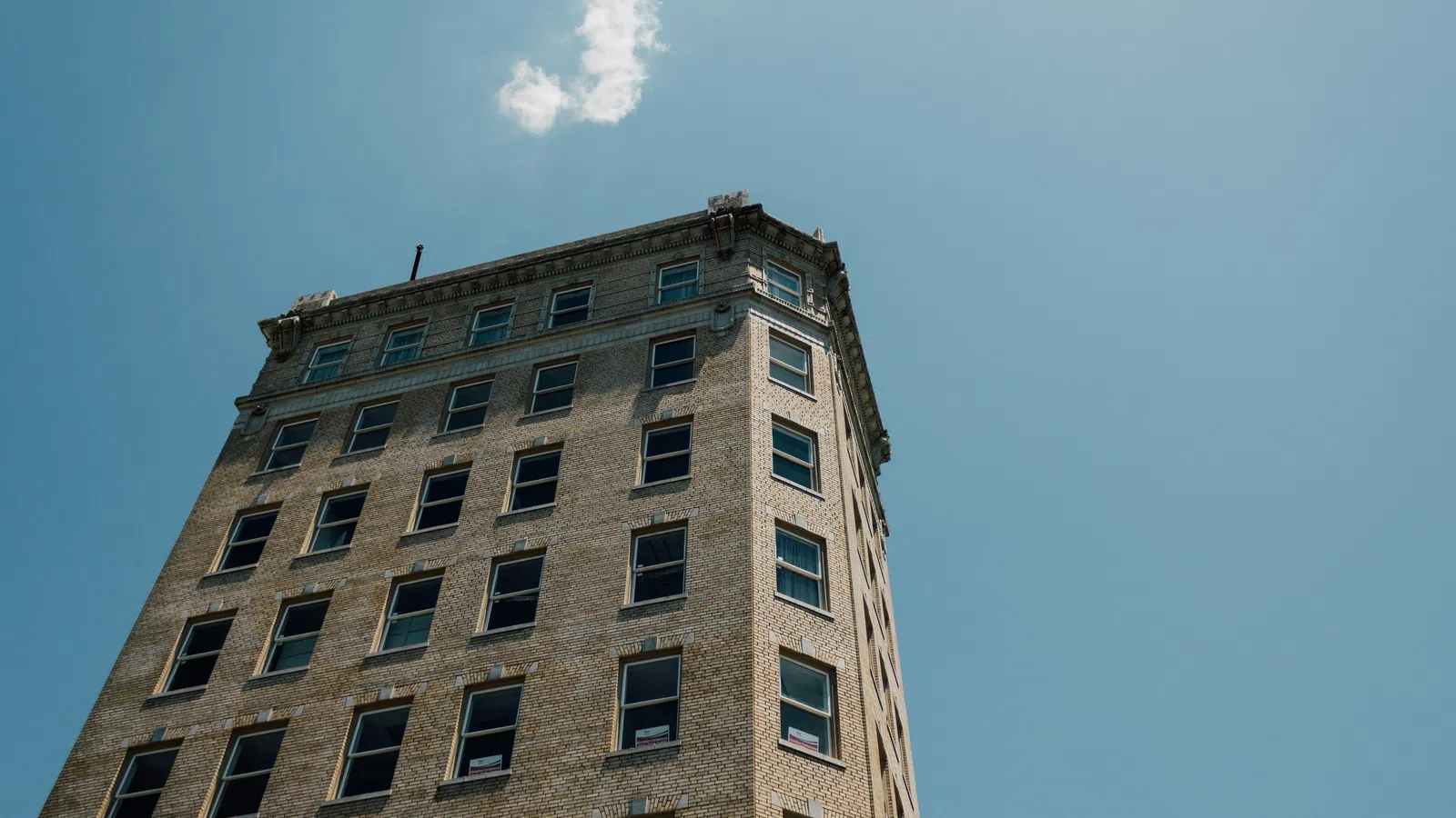 Tall historic brick building with many windows under a clear blue sky with a small cloud of white smoke.