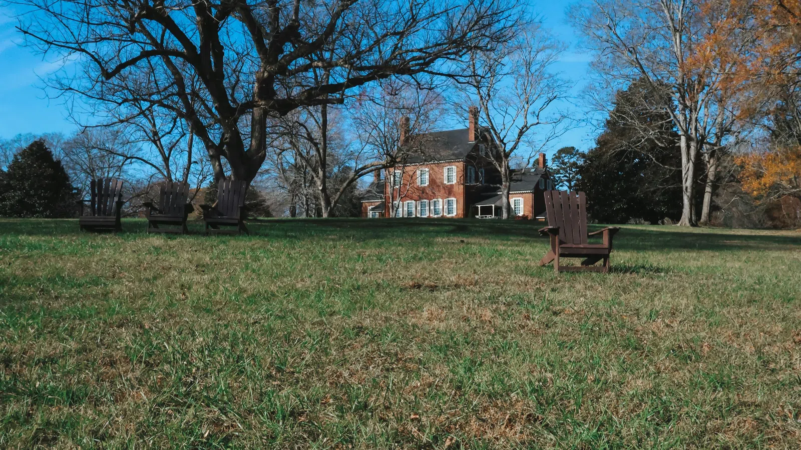 Historic brick house with white shutters on a grassy hill under clear blue sky with leafless and autumn trees.