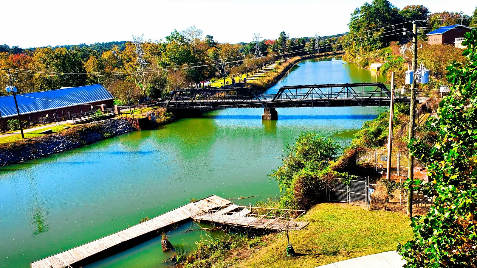 Calm river with a metal bridge, wooden dock, green trees, and buildings under a clear sky