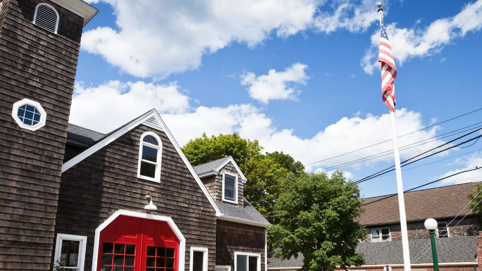 Historic wooden firehouse with red doors, American flag, power lines, and blue sky with clouds in small town.