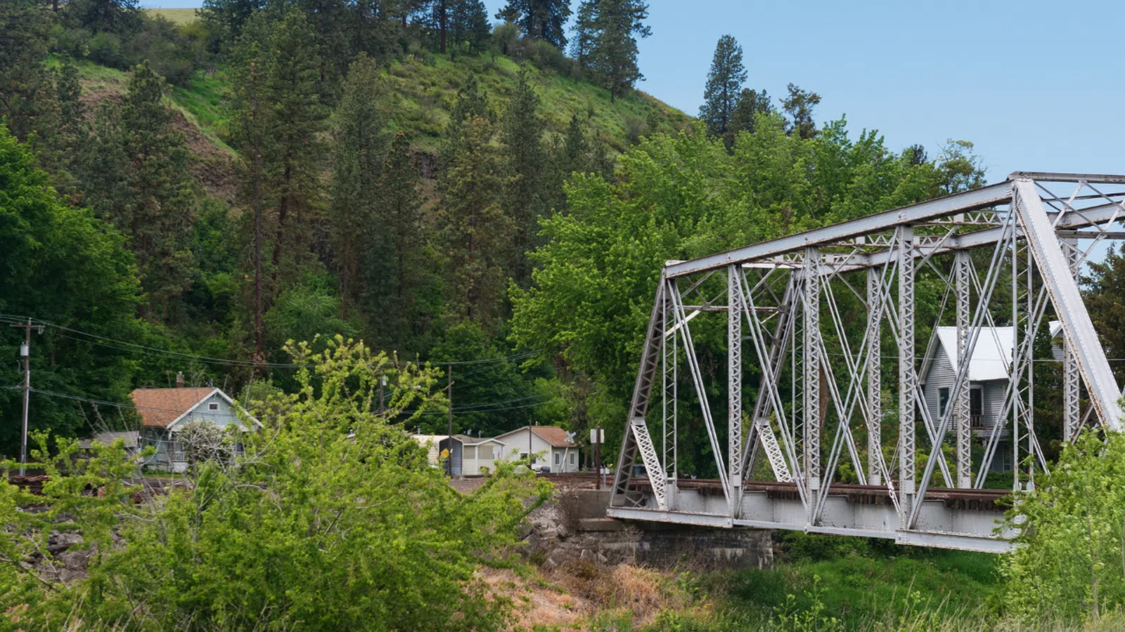 Rustic white metal truss bridge over river surrounded by lush greenery and forested hillside on a clear day