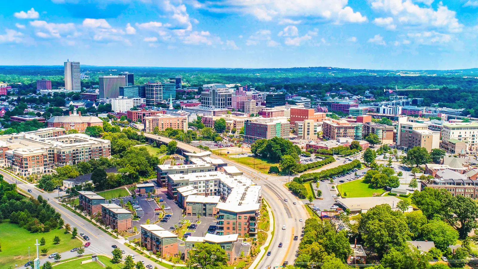 Aerial view of a modern cityscape with roads, buildings, and lush green trees under a clear sky.
