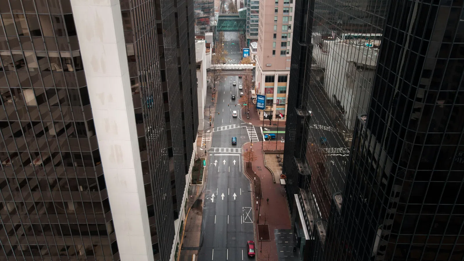 Aerial view of a city street flanked by tall glass skyscrapers with cars and pedestrians in an urban setting.
