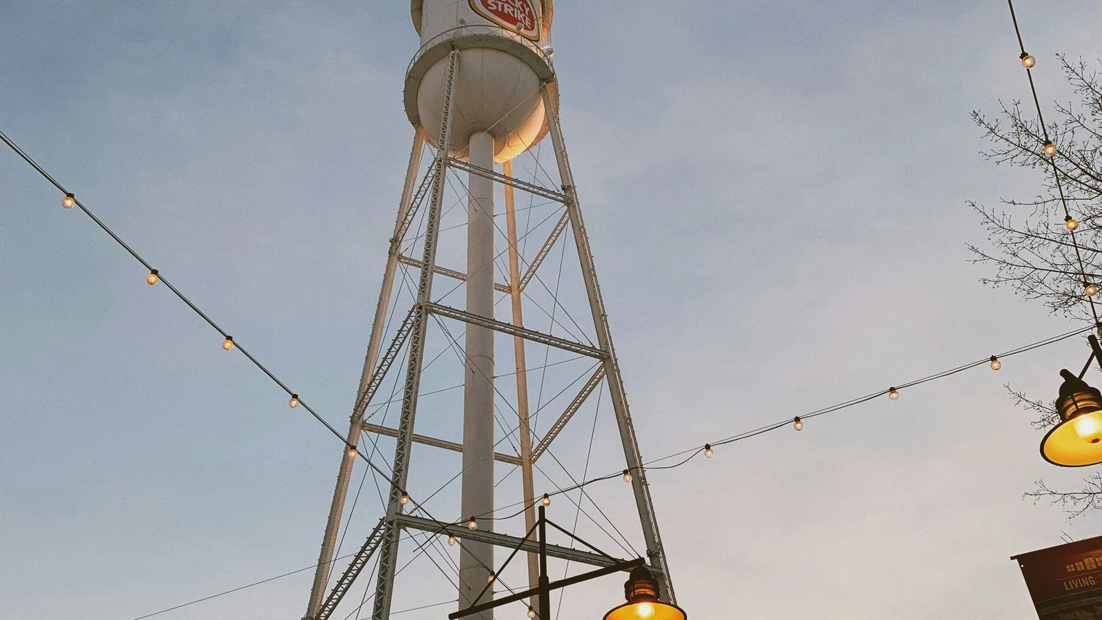Tall Lucky Strike water tower at dusk with string lights and street lamps glowing under a clear sky.