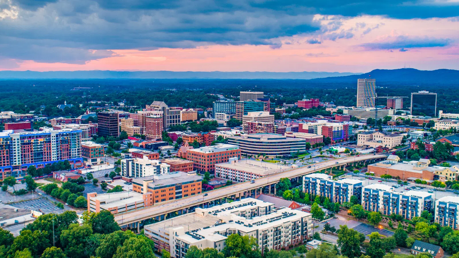 Aerial view of a city skyline at sunset with buildings, roads, and greenery under a cloudy sky.