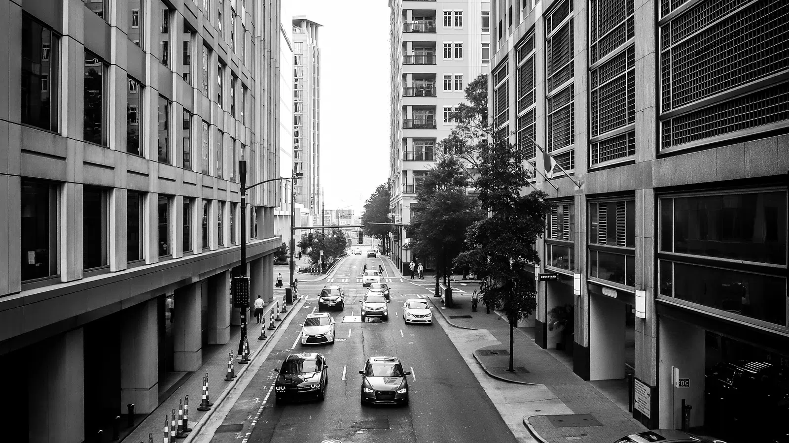 Black and white city street scene with cars driving between tall office buildings on an urban road.