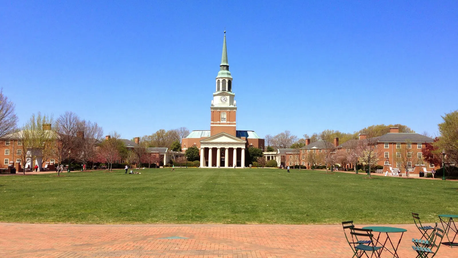 Green lawn and brick plaza with tables in front of a central building with clock tower and steeple under clear blue sky