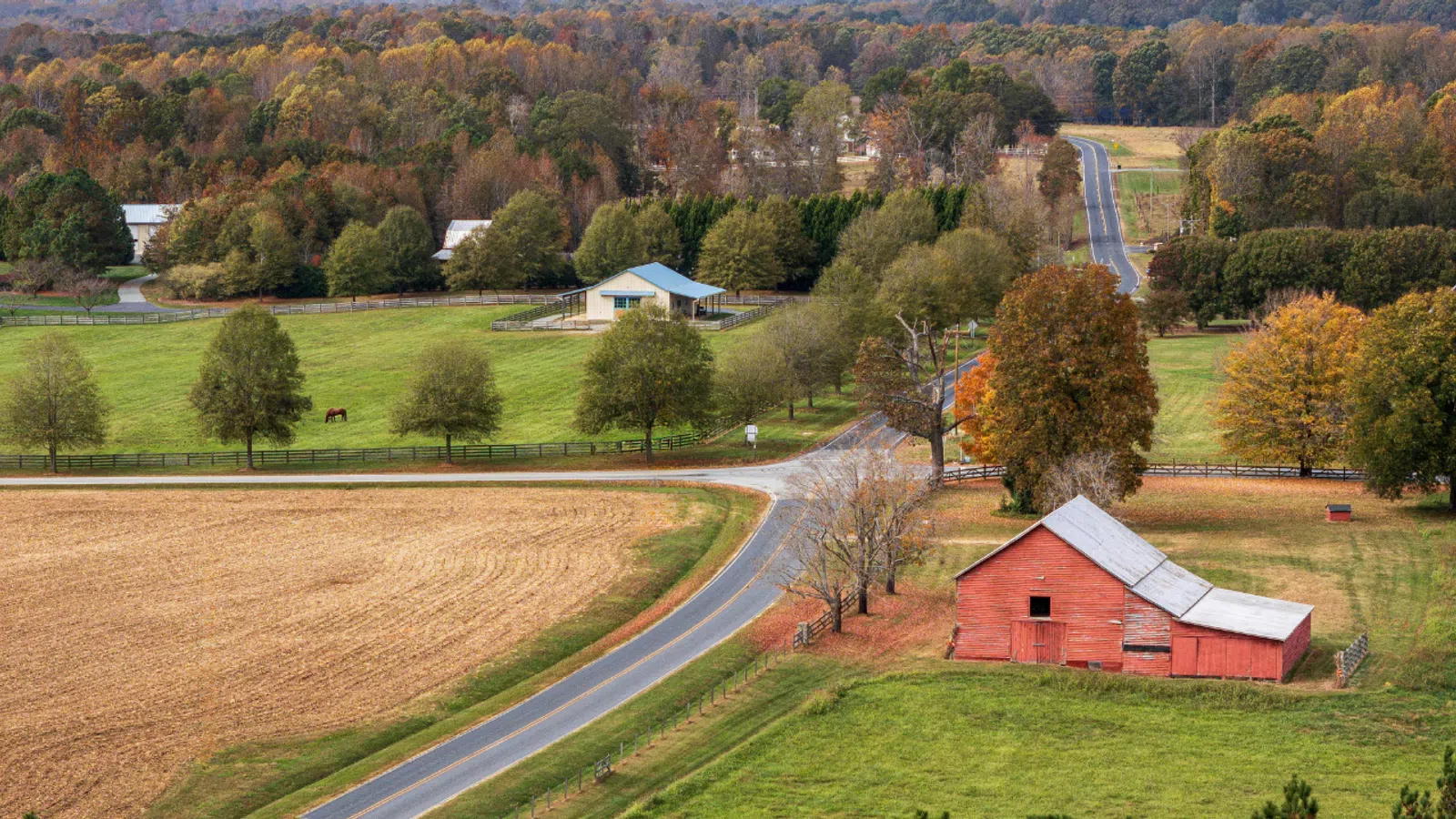 Aerial view of a rural landscape with a red barn, winding road, fields, trees, and autumn colors.