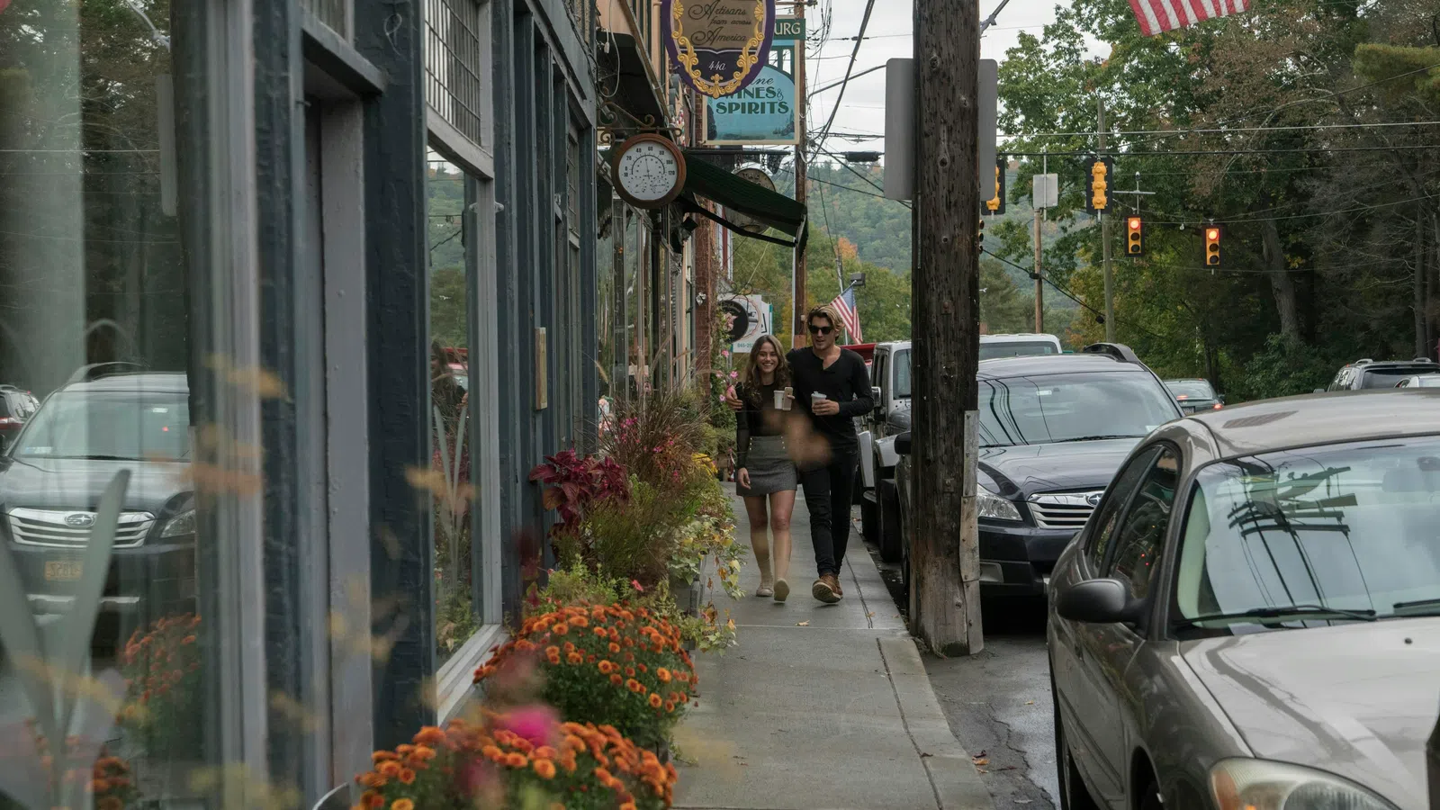 Couple walking and holding coffee cups on a sidewalk with flowers, parked cars, and American flags in a Selma.