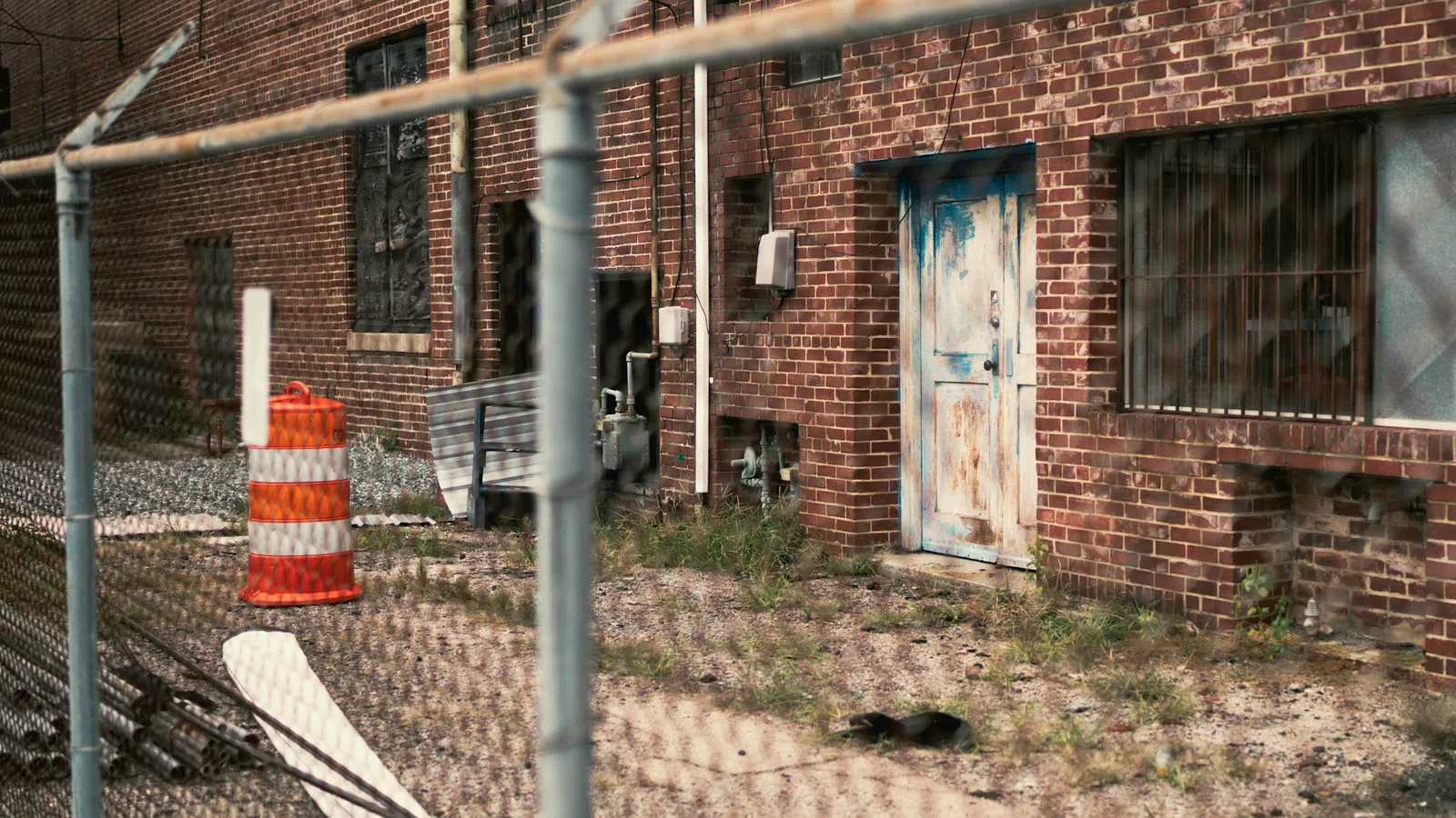 Rusty white door and barred windows on brick building behind chain-link fence with construction barrel