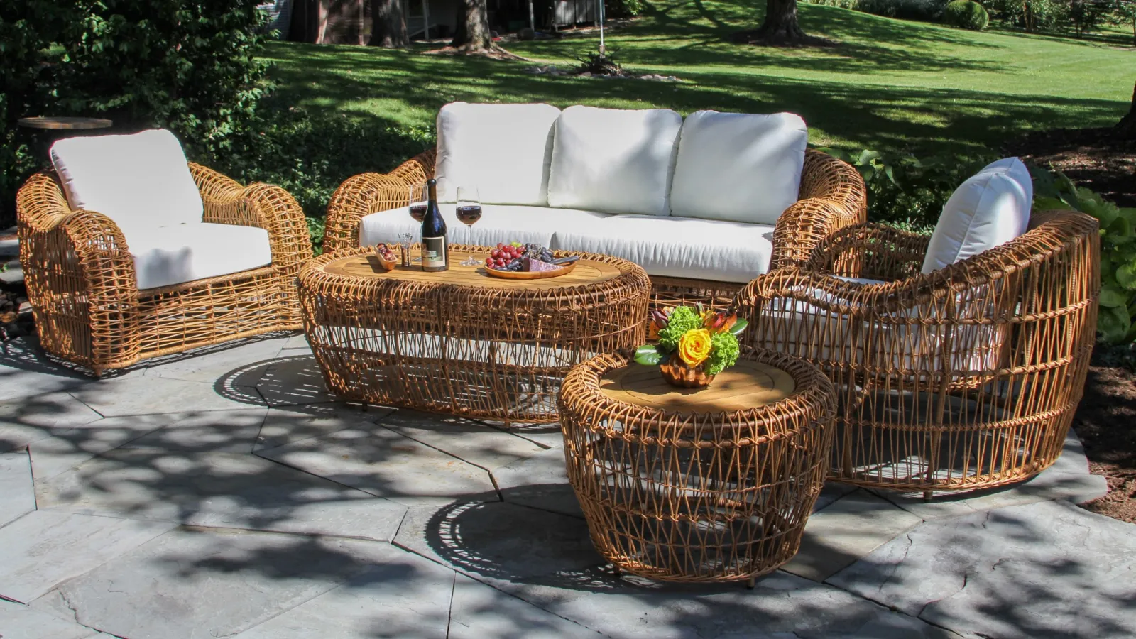 Outdoor wicker patio furniture set with white cushions on stone patio in sunny backyard.