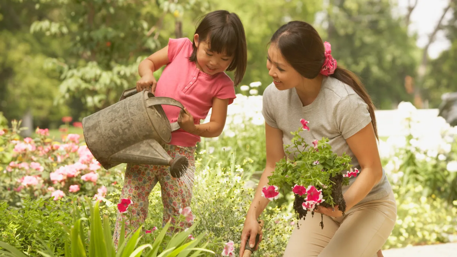 Mother and daughter gardening together with watering can and flowers in a bright outdoor garden.