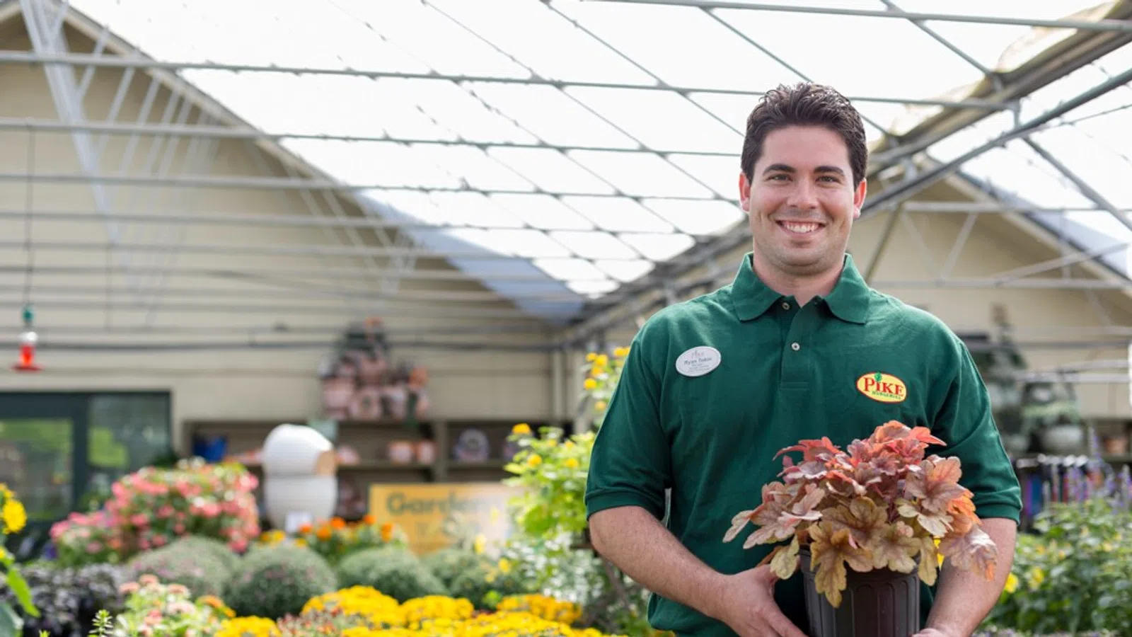 Smiling gardener in green polo holding potted plant inside greenhouse with colorful flowers in background