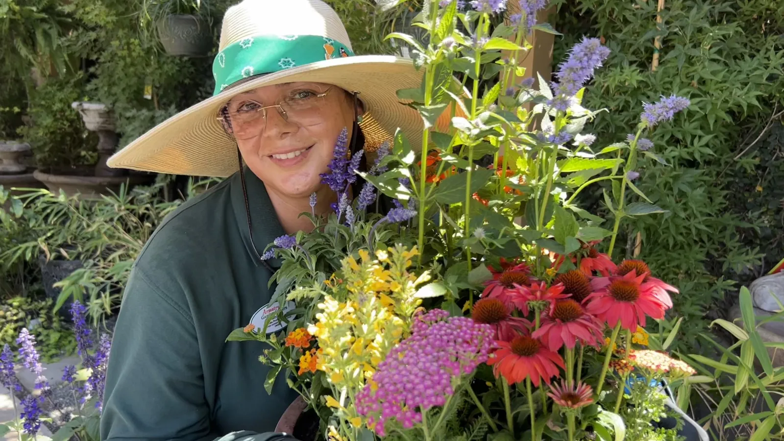 Smiling gardener wearing wide-brimmed hat holding a colorful bouquet of mixed flowers in a lush garden.