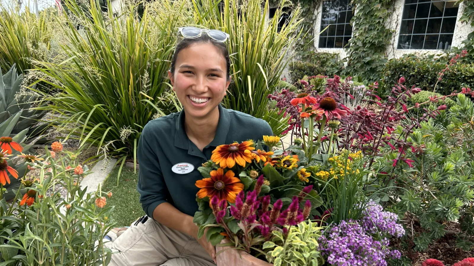 Smiling gardener arranging vibrant flowerpot with sunflowers, purple and pink blooms in lush garden setting.