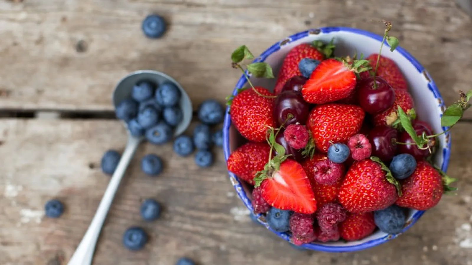 Bowl of fresh strawberries, cherries, raspberries, and blueberries with spoonful of blueberries on rustic wooden table.