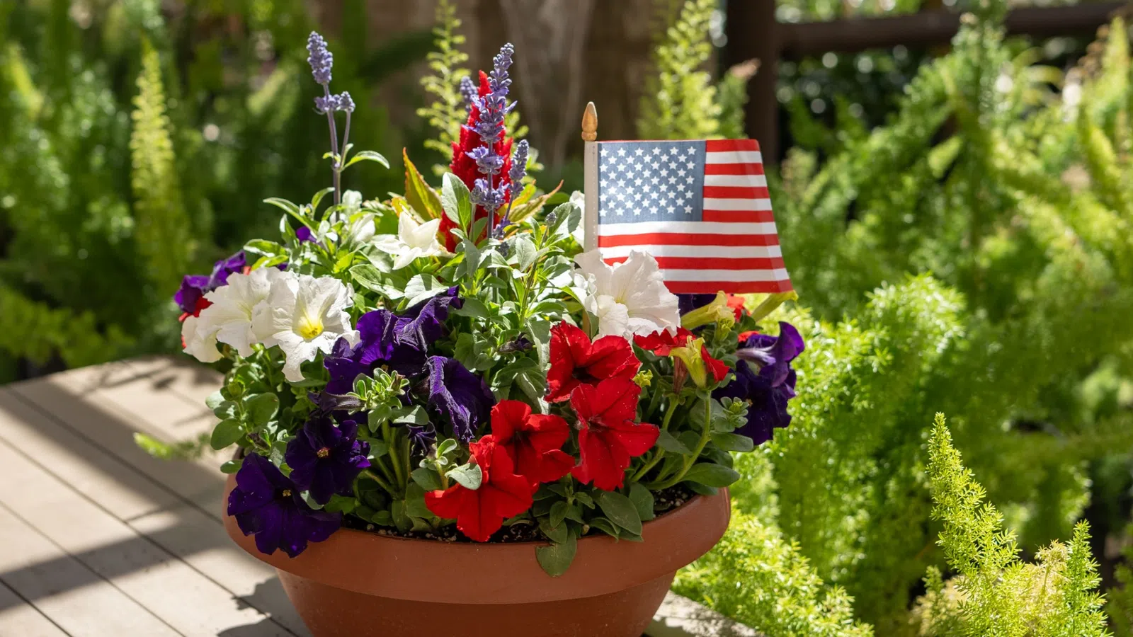 Potted flowers in red, white, and purple with a small American flag on a wooden table outdoors.
