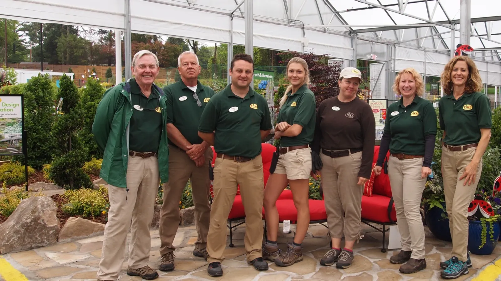 Seven nursery staff members in green shirts posing inside a greenhouse with plants and outdoor furniture.
