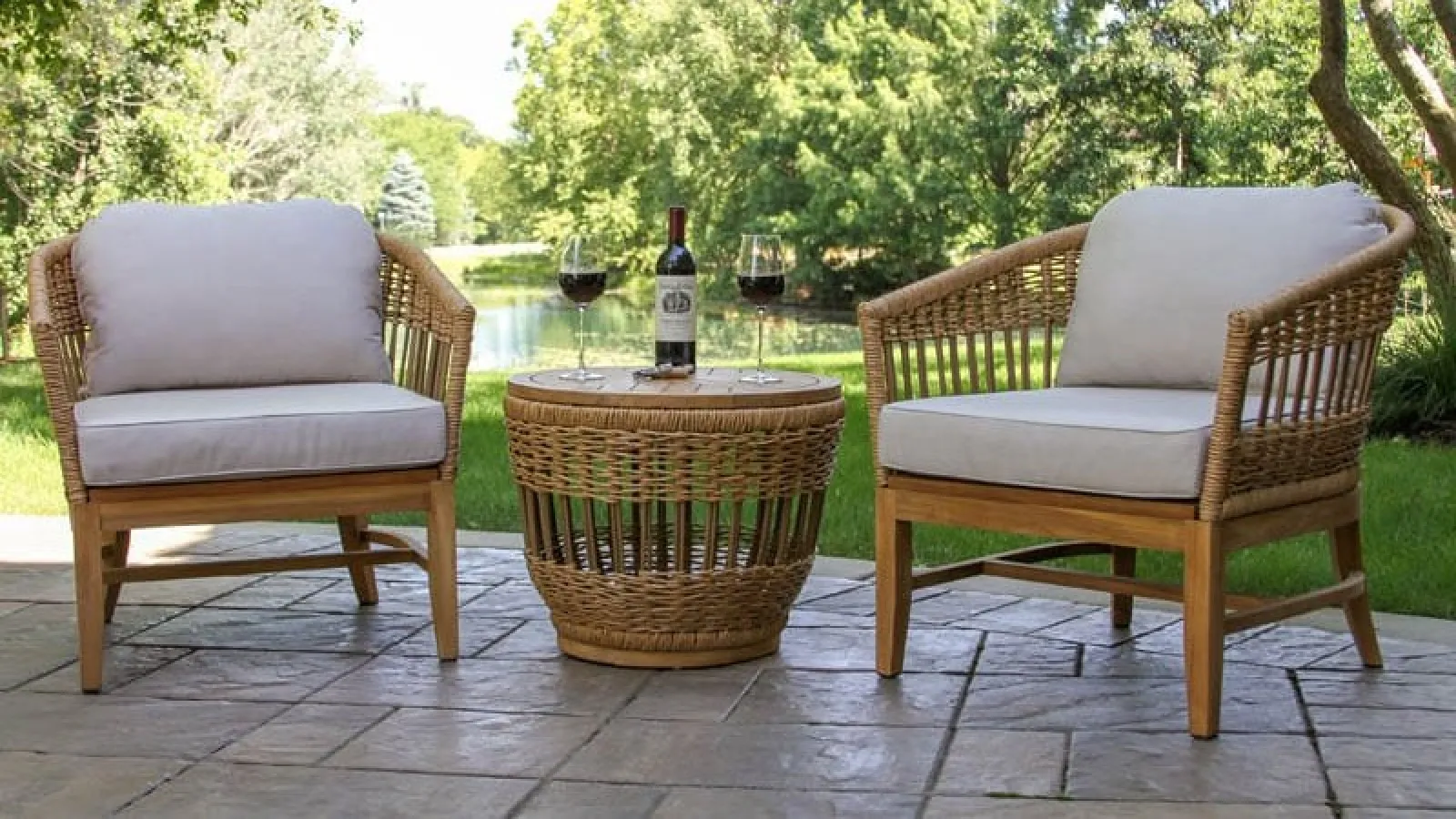 Two wicker armchairs with cushions and a matching round table with wine glasses and bottle on a stone patio