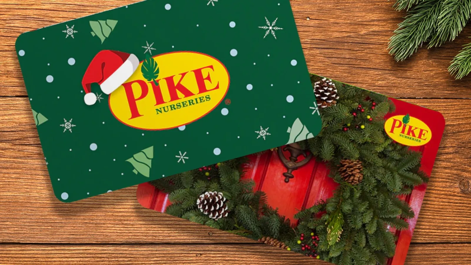 Two Pike Nurseries gift cards with Christmas themes on a wooden table beside pine branches and a red ornament