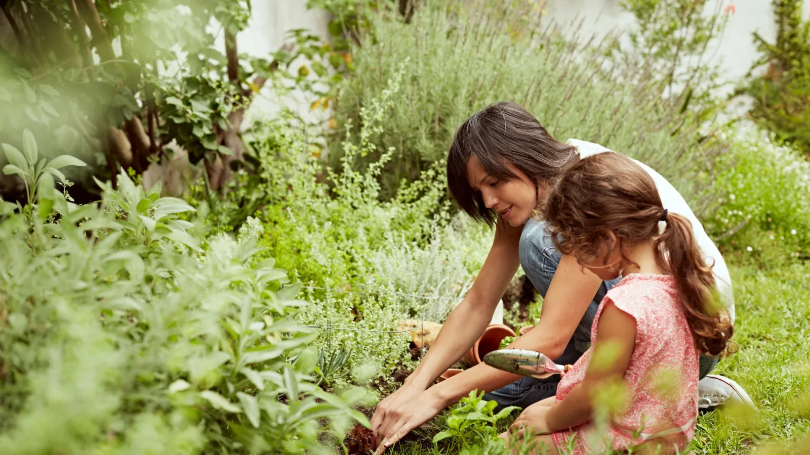 Mother and daughter gardening together in a lush green backyard filled with herbs and plants.