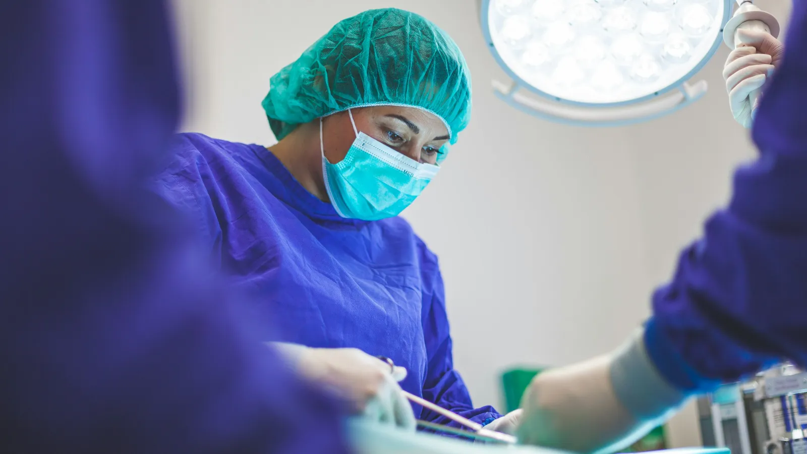 Surgeon wearing surgical mask and cap performing operation under bright operating room lights.