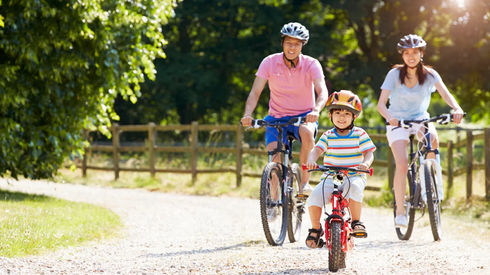 Family of three wearing helmets riding bikes on a sunny dirt path surrounded by trees and wooden fence.