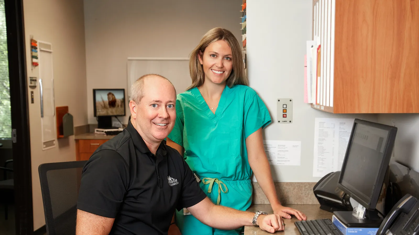 Veterinarian in black shirt and vet nurse in green scrubs smiling at a desk in a veterinary clinic office.