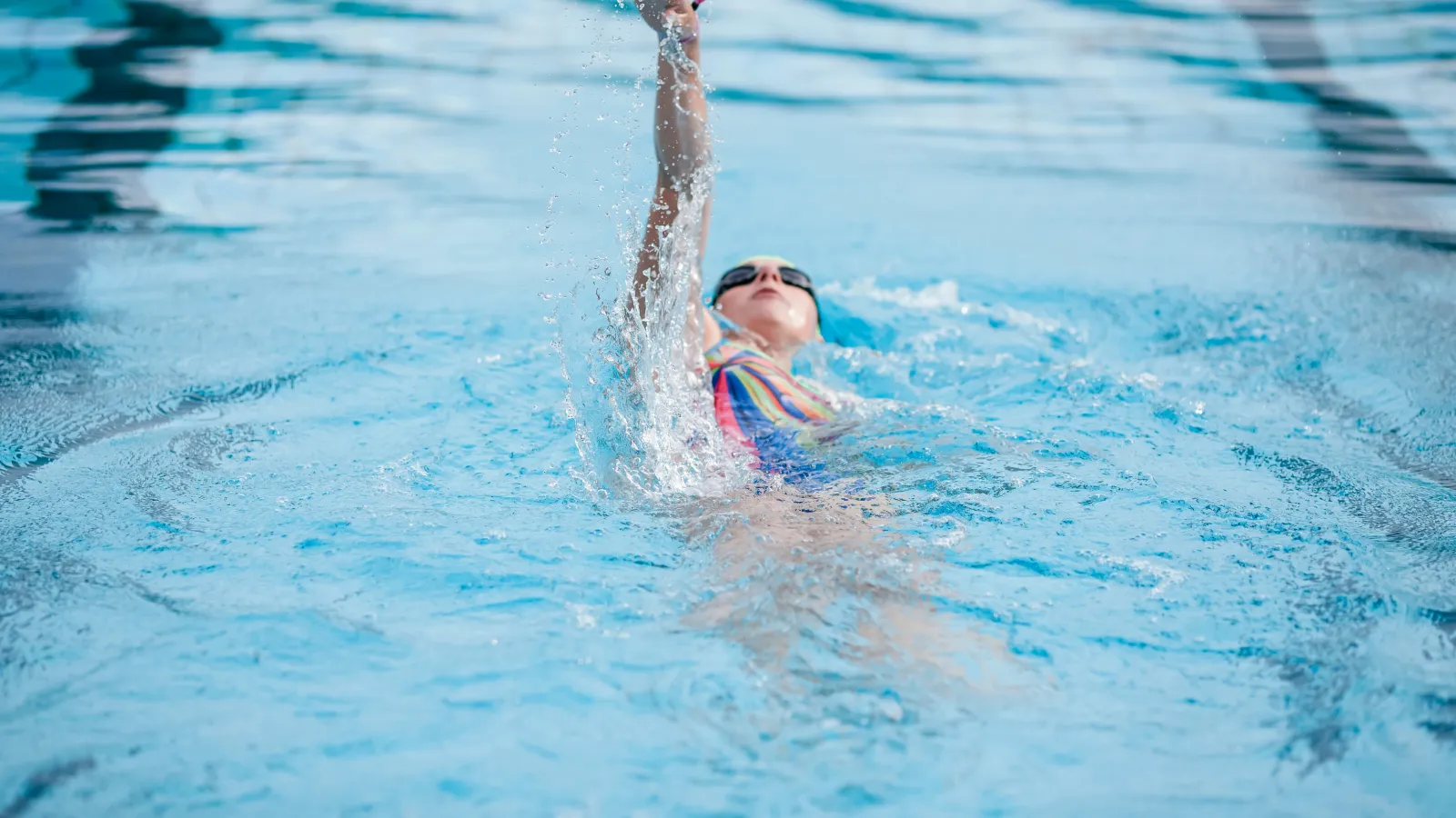 Swimmer in colorful swimsuit performs backstroke in clear blue pool water with splash effect.
