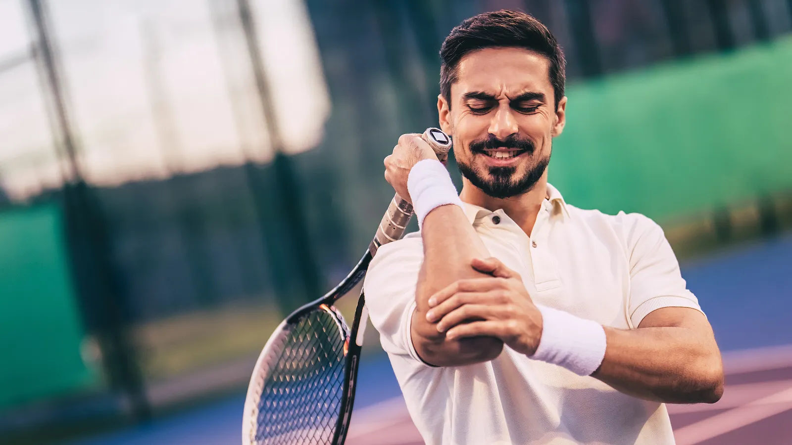 Male tennis player in white shirt holding his elbow in pain on an outdoor tennis court with racket.