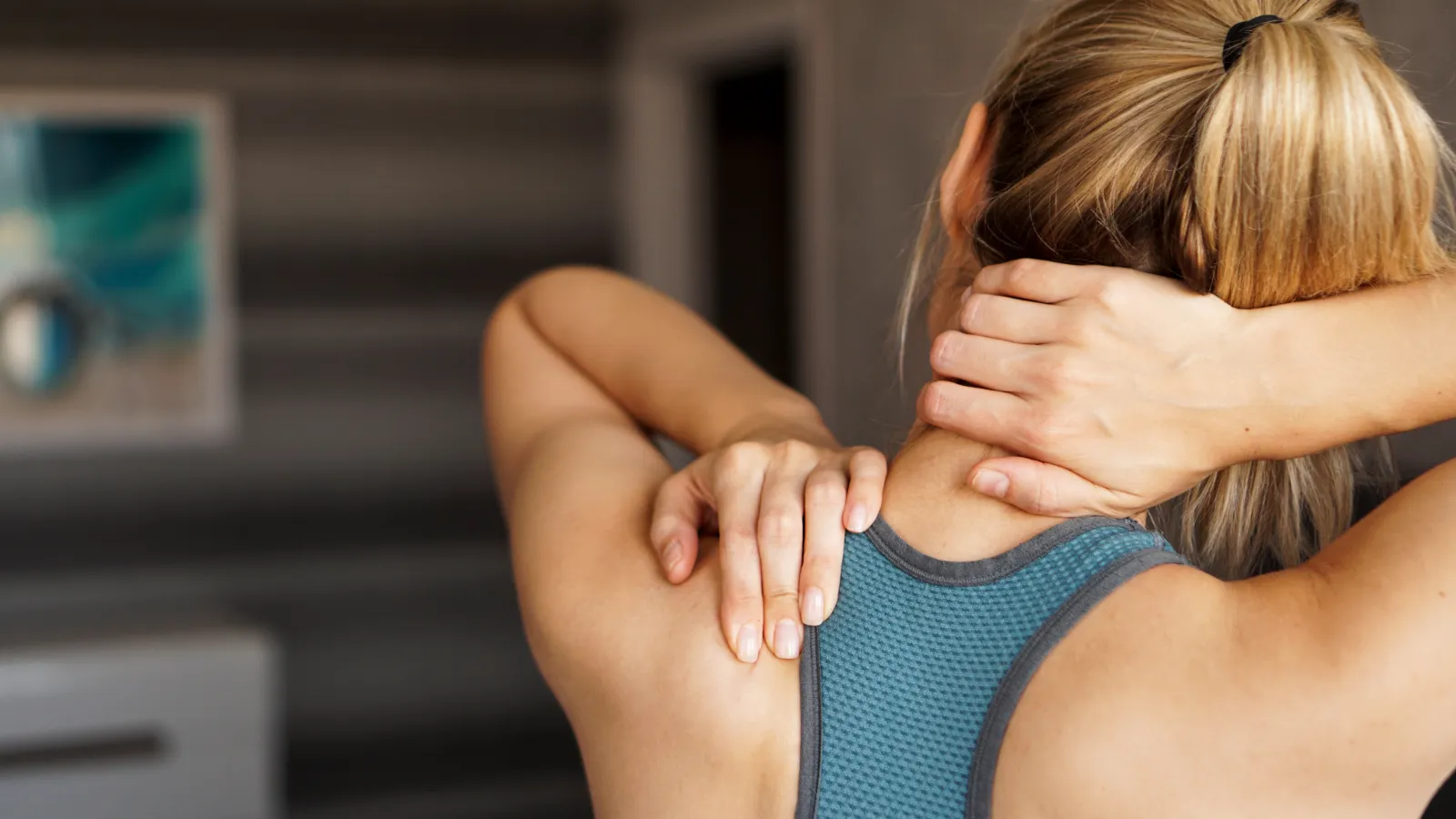 Woman in sportswear massaging her neck and upper back indoors, showing muscle tension relief.