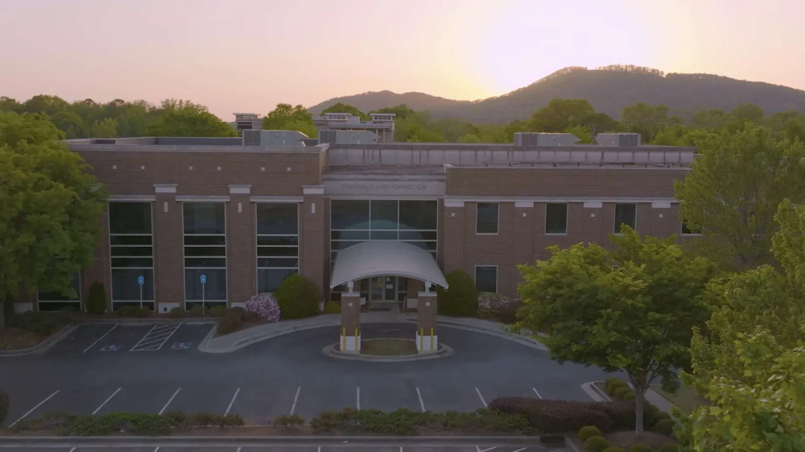 Modern brick office building with large windows at sunset, surrounded by trees and mountains in the background.
