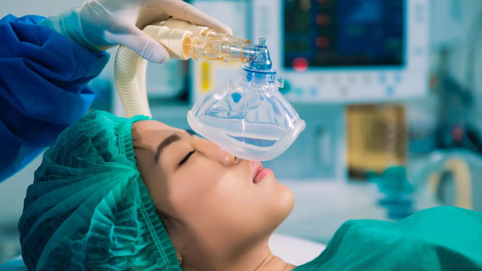 Patient in operating room receiving anesthesia through a mask held by a gloved medical professional.