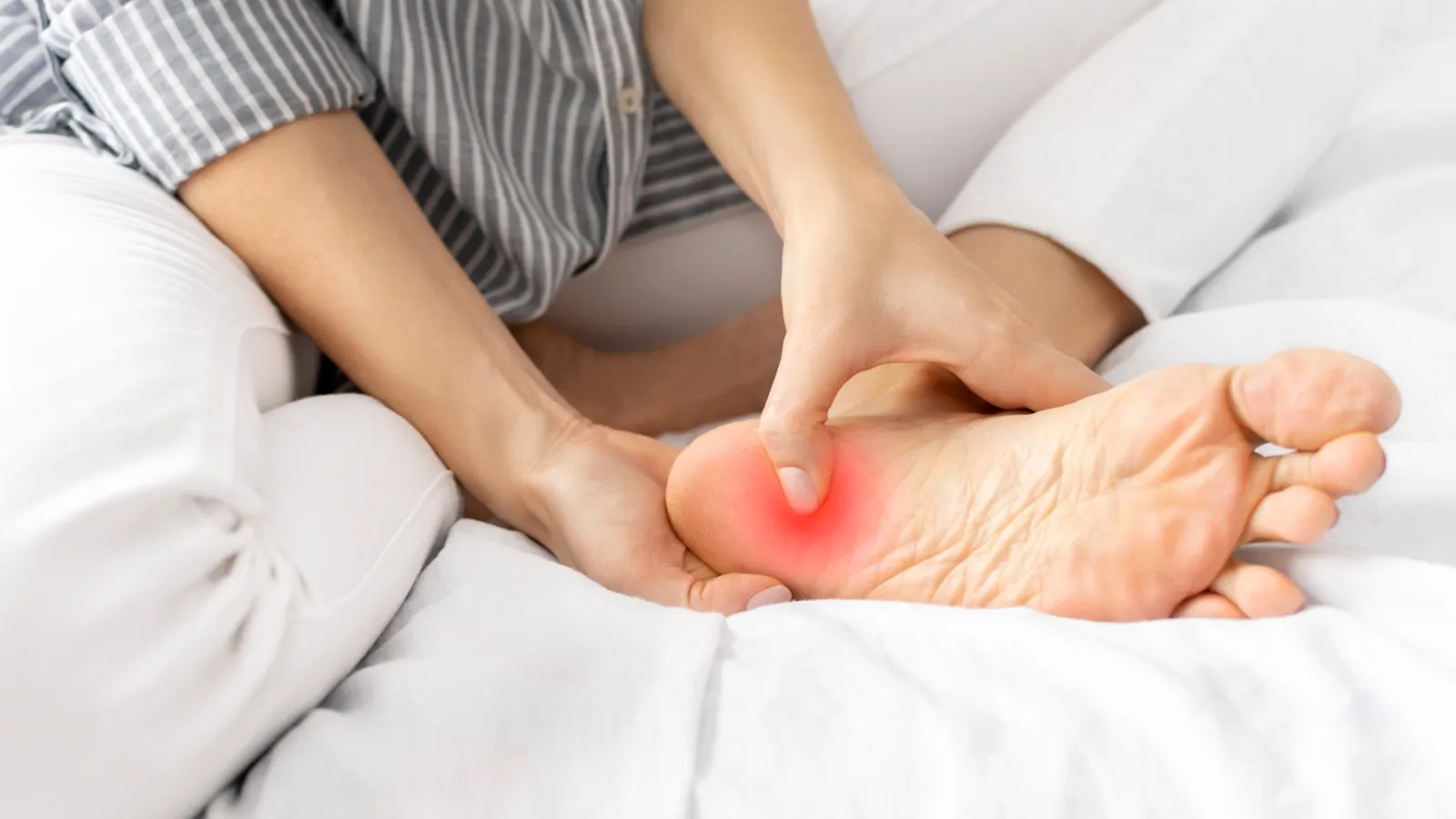 Person rubbing the sole of their foot highlighting pain area in red on a white bed.